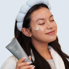 Woman applying cream to her face with a blue towel on her head, holding a skincare product.